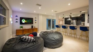 A modern living room with a large flat-screen TV, bean bags, a kitchen area with bar stools, and a sliding glass door leading to an outdoor space. - Villa Azur by Glam Homes (North Miami Beach)