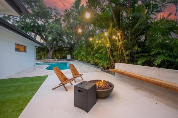 Eine moderne Terrassenanlage im Freien mit Pool, zwei Holzstühlen, einer Feuerstelle und Lichterketten.