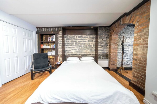 A bedroom with a bed, a chair, a bookshelf, and a brick wall.