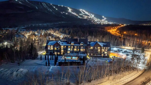 Un grand bâtiment à plusieurs étages, dont le toit est recouvert de neige, entouré d’une zone boisée et avec une montagne en arrière-plan.