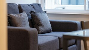 A grey sofa with cushions, a wooden floor, and a window with a view of a building. - 8 Stanley Street (Liverpool)