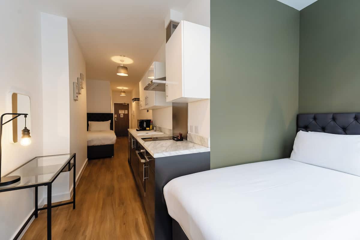 Une chambre d’hôtel moderne équipée d’un lit, d’une petite kitchenette, d’une table en verre et d’un couloir menant à la salle de bain.