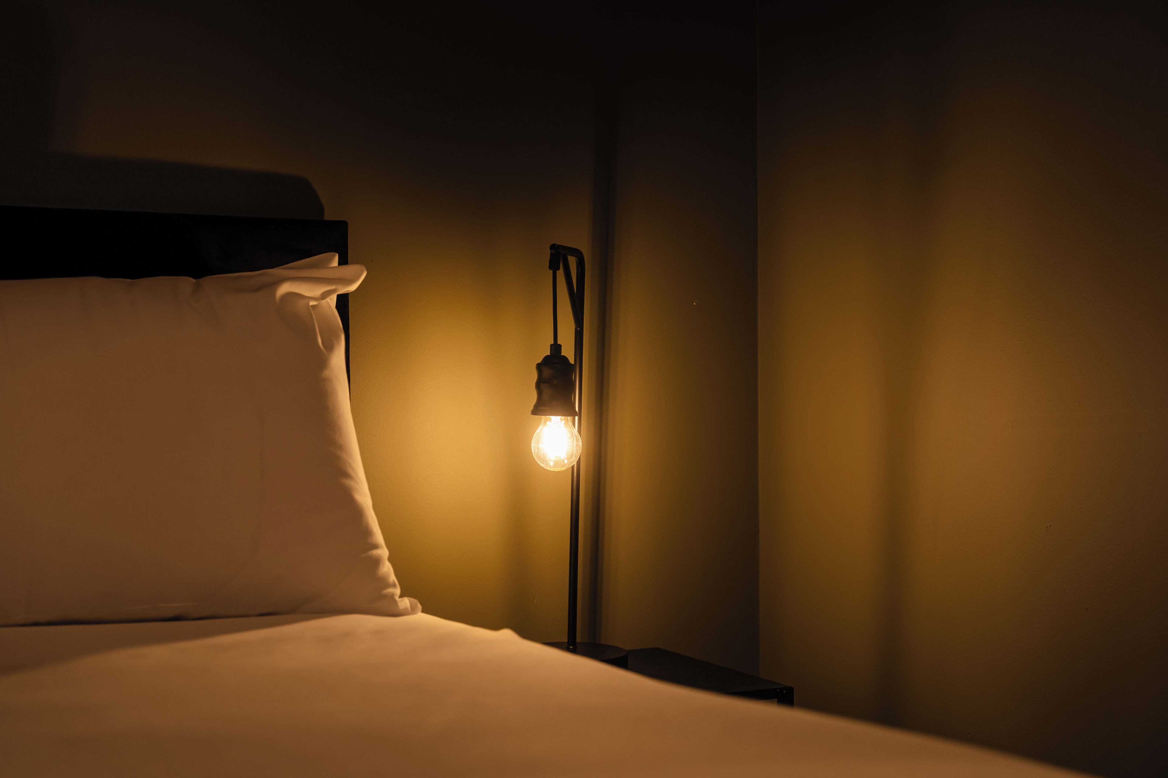 A bed with white bedding, a nightstand with a lamp, and a wall with a curtain.