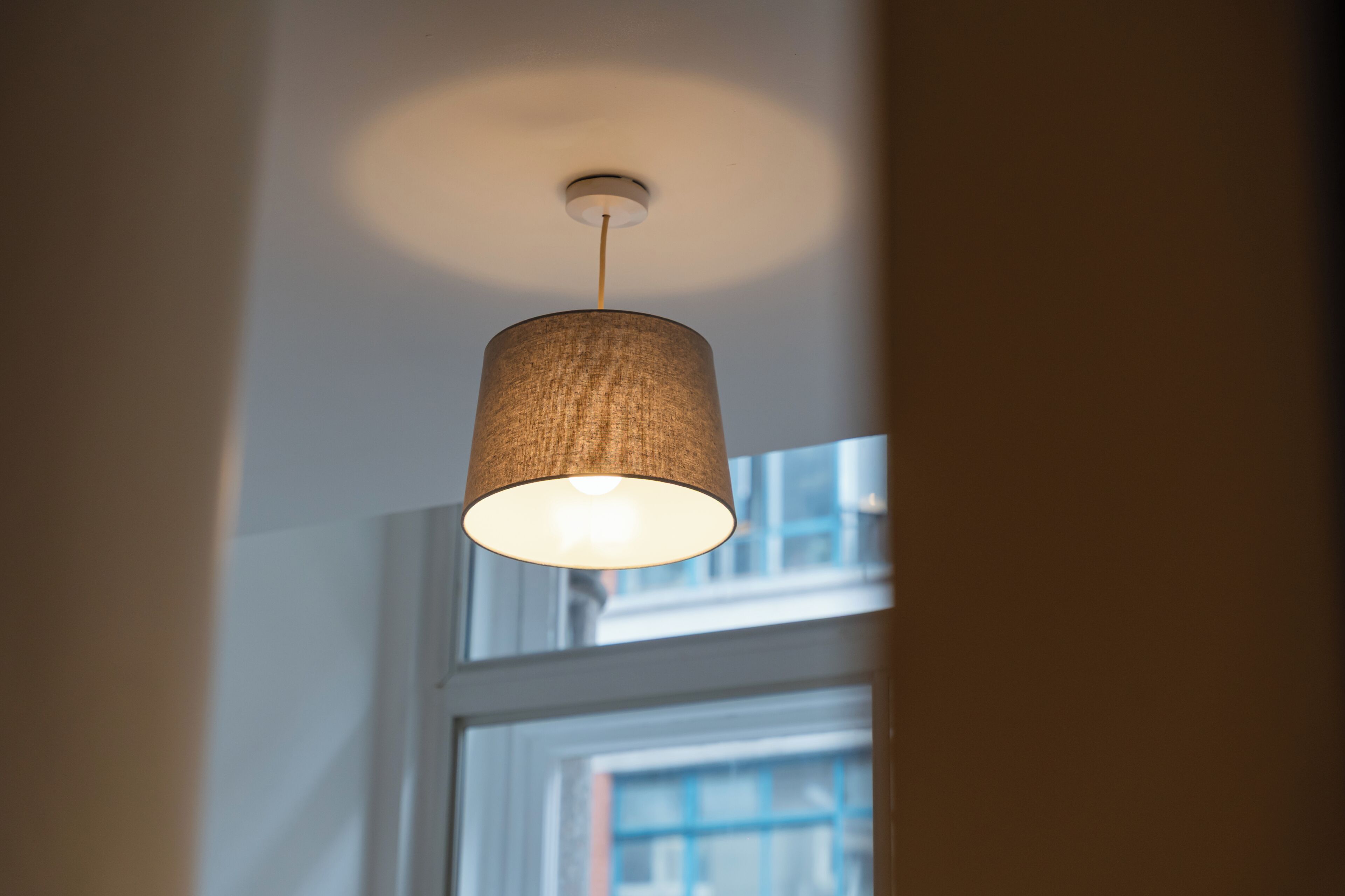 Une lampe suspendue avec un abat-jour cylindrique, placée dans une pièce dotée d’une fenêtre.