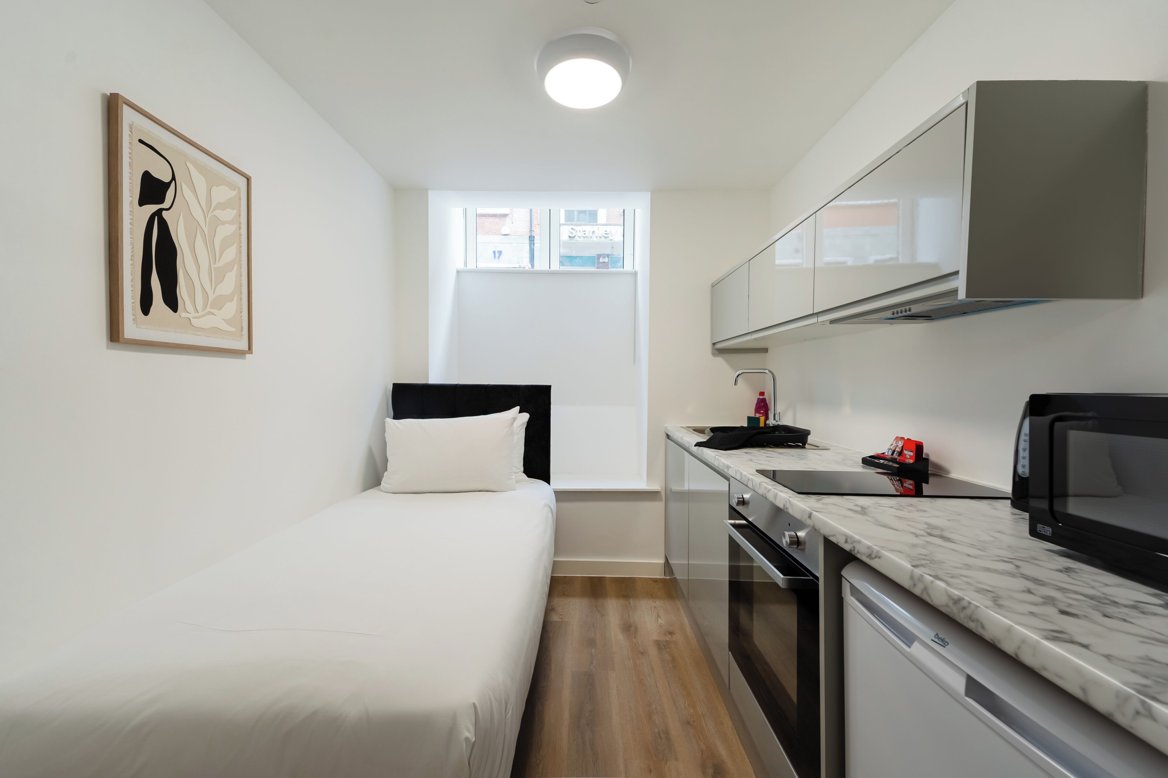 A compact room with a bed, kitchenette, and a window with a view of buildings.