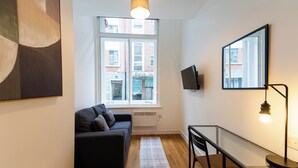A modern living room with a sofa, a dining table, a TV mounted on the wall, and a large window with a view of buildings. - 8 Stanley Street (Liverpool)