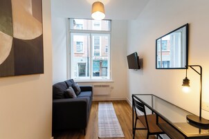 Una sala de estar moderna con un sofá, una mesa de comedor, un televisor montado en la pared y un ventanal con vistas a edificios.