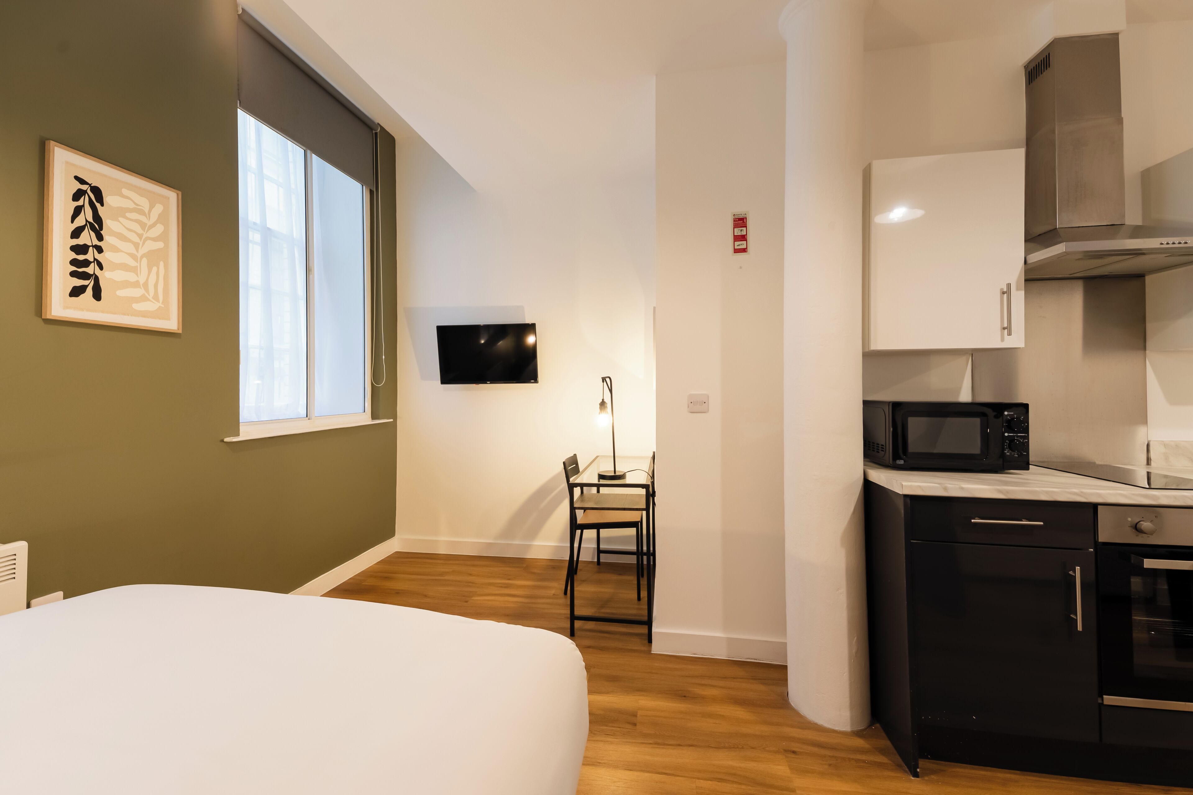 Een kleine hotelkamer met een bed, een bureau met stoel, een televisie, een magnetron en een kitchenette.