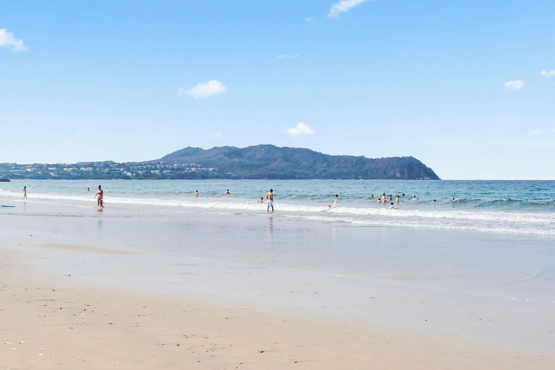 Una playa con gente nadando y caminando, una montaña al fondo y un cielo despejado.