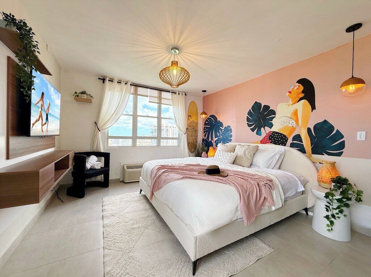 Une chambre à coucher comprenant un lit, un téléviseur, une fenêtre avec des rideaux, une fresque murale et un luminaire au plafond.