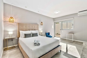 Ein modernes Schlafzimmer mit einem großen Bett, Nachttischlampen, einem blauen Sessel und Blick ins Freie.