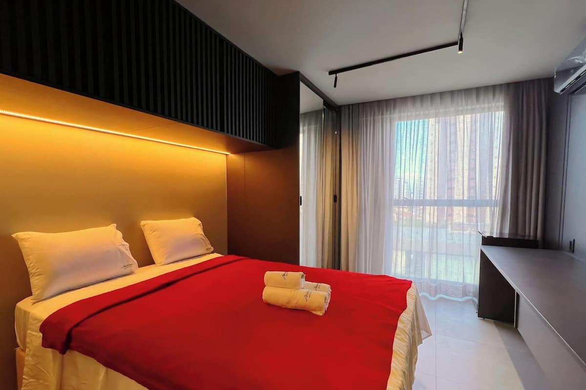Una habitación de hotel con cama, una colcha roja, almohadas blancas, escritorio y ventana con cortinas.