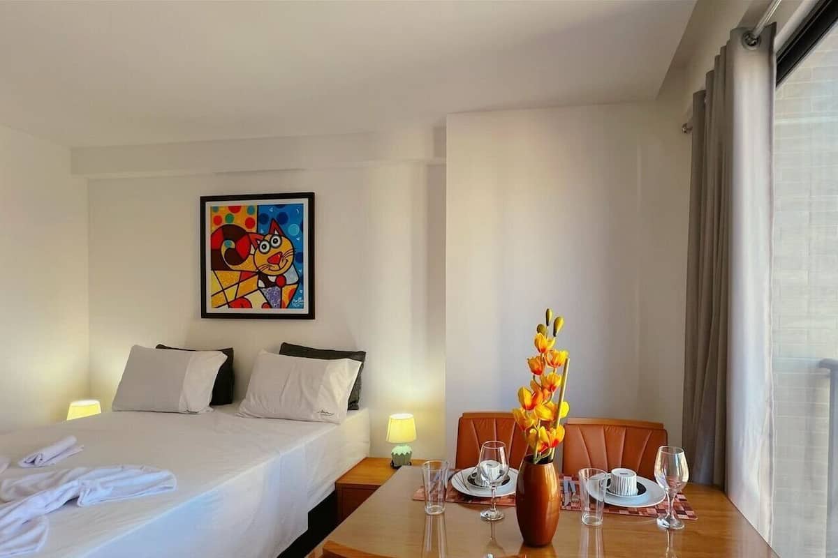Habitación de hotel con una cama, una mesa de comedor dispuesta con platos, vasos y un florero.