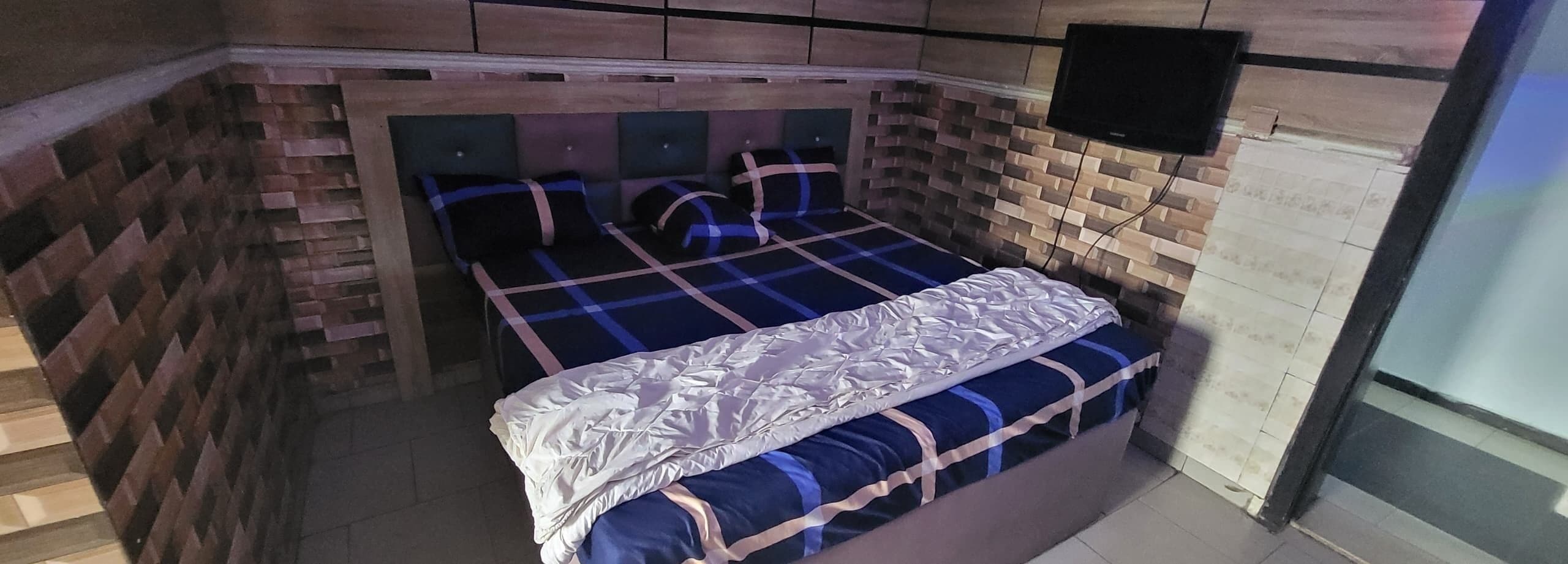 Una habitació amb un llit, una televisió penjada a la paret i una paret amb relleu de maó.