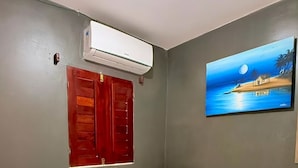 A single bed with a white bedspread, a red door, an air conditioning unit, a small white refrigerator, and a painting of a beach scene on the wall. - Pousada Anauá - Jericoacoara (Jijoca de Jericoacoara)