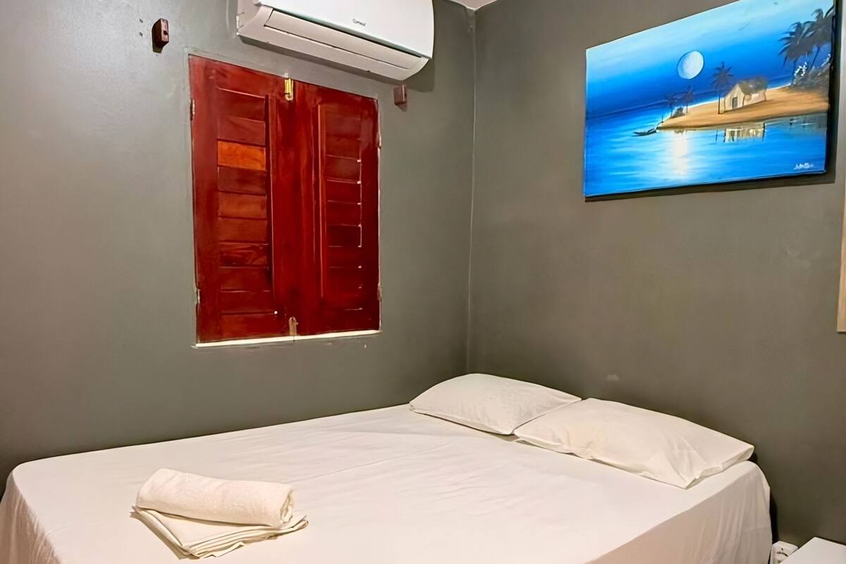 A single bed with a white bedspread, a red door, an air conditioning unit, a small white refrigerator, and a painting of a beach scene on the wall.