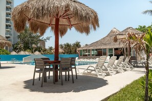 A poolside area with lounge chairs, a table, and a thatched umbrella. - Ocean View Studio Beachfront in Hotel Zone (Cancun)