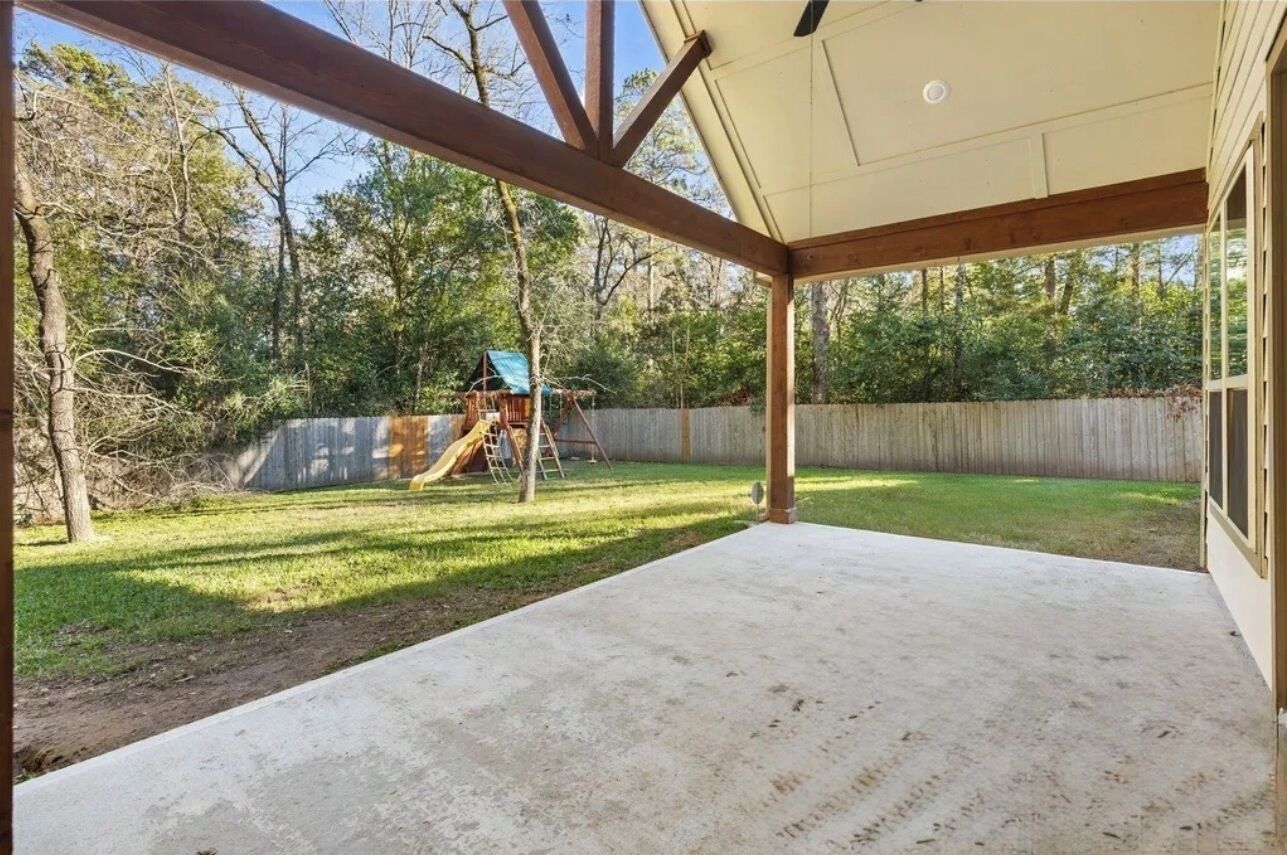 Ein überdachter Patio-Bereich mit Betonboden, Holzbalken und Blick auf einen Garten mit Rutsche und Spielgeräten.