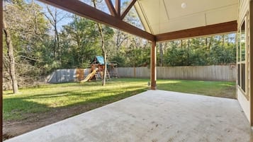 Ein überdachter Patio-Bereich mit Betonboden, Holzbalken und Blick auf einen Garten mit Rutsche und Spielgeräten.