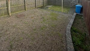 Un jardin à l’arrière d’une maison, avec une zone recouverte de gravier, une clôture en bois et des maisons en arrière-plan.
