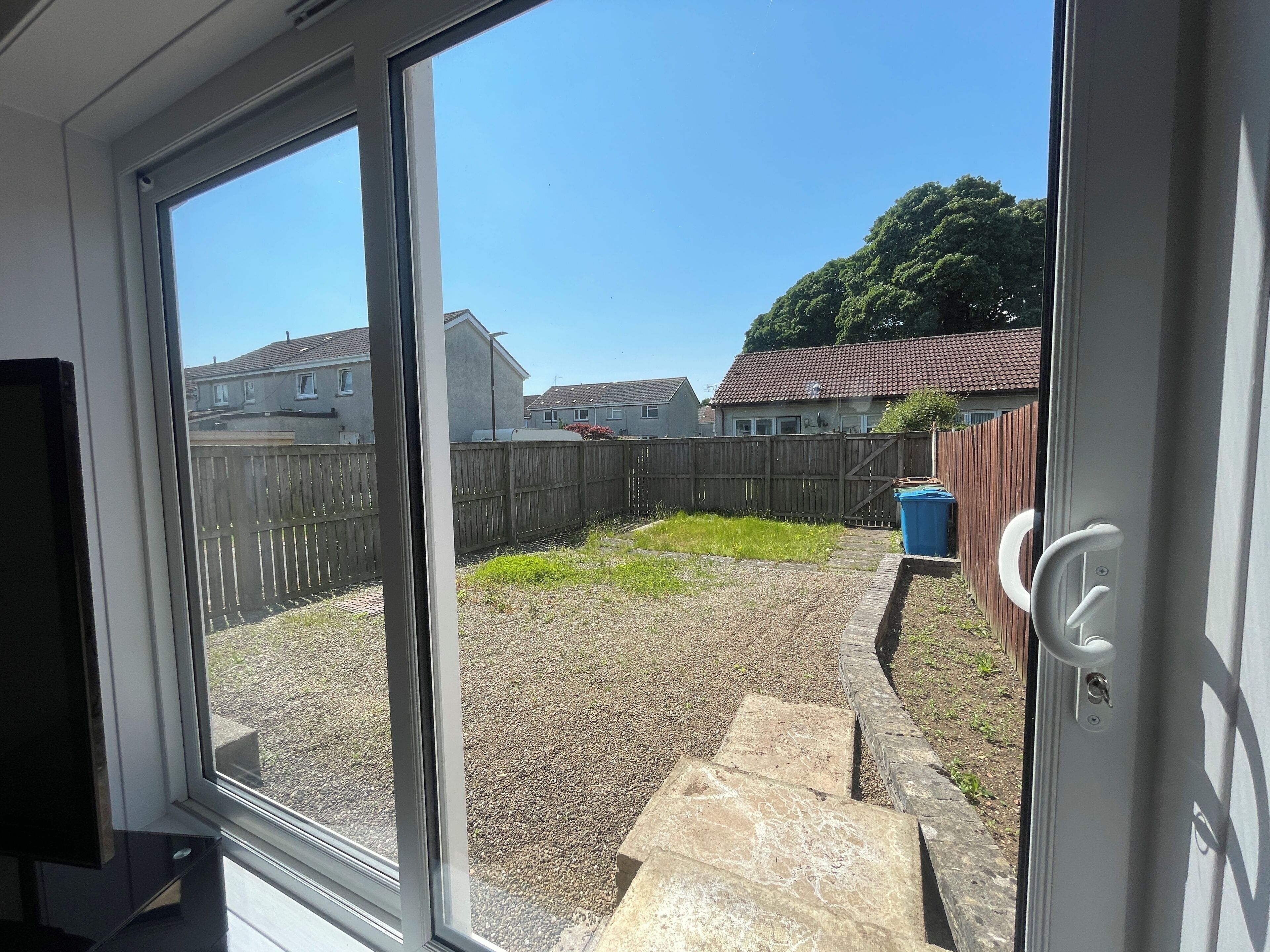 A view through a window of a backyard with a gravel path, a wooden fence, and a blue trash bin.