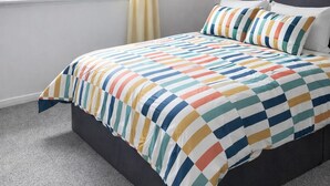 Une chambre moderne avec un mur à rayures, un lit recouvert d’une courtepointe colorée à rayures, une fenêtre avec un rideau et un luminaire au plafond.