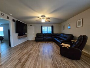 Un salon spacieux comprenant un canapé d’angle, un fauteuil inclinable, un ventilateur de plafond et un téléviseur à écran plat fixé au mur.