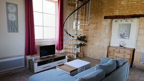 A living room with a blue sofa, a TV on a wooden stand, a staircase, and a stone wall. - "La Borie" charming gîte with swimming pool in Daglan at Mas de Cause (Daglan)