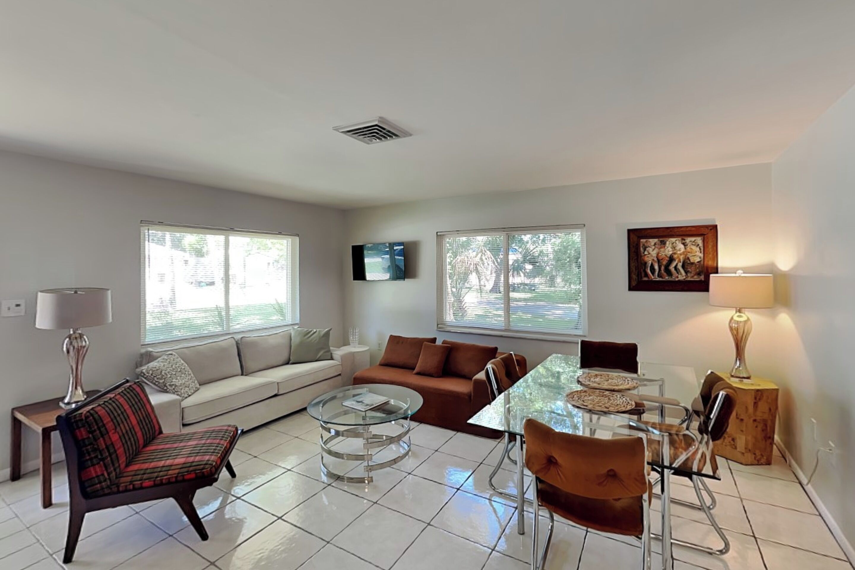 A living room with a sofa, armchairs, a glass coffee table, a dining table with chairs, and a TV mounted on the wall.