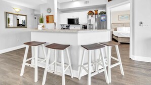A modern kitchen with white cabinetry, a central island with stools, a microwave, refrigerator, and a coffee maker. - Celadon 409- Champagne Sunsets (Panama City Beach)