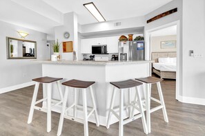 A modern kitchen with white cabinetry, a central island with stools, a microwave, refrigerator, and a coffee maker. - Celadon 409- Champagne Sunsets (Panama City Beach)