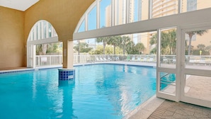 An indoor swimming pool with clear blue water, large arched windows, and a view of palm trees and buildings outside. - Celadon 409- Champagne Sunsets (Panama City Beach)