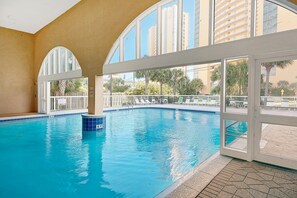An indoor swimming pool with clear blue water, large arched windows, and a view of palm trees and buildings outside. - Celadon 409- Champagne Sunsets (Panama City Beach)