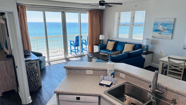 A modern kitchen with a Bosch dishwasher, a sink, and a dining area with a blue sofa and a view of the ocean. - Celadon 409- Champagne Sunsets (Panama City Beach)