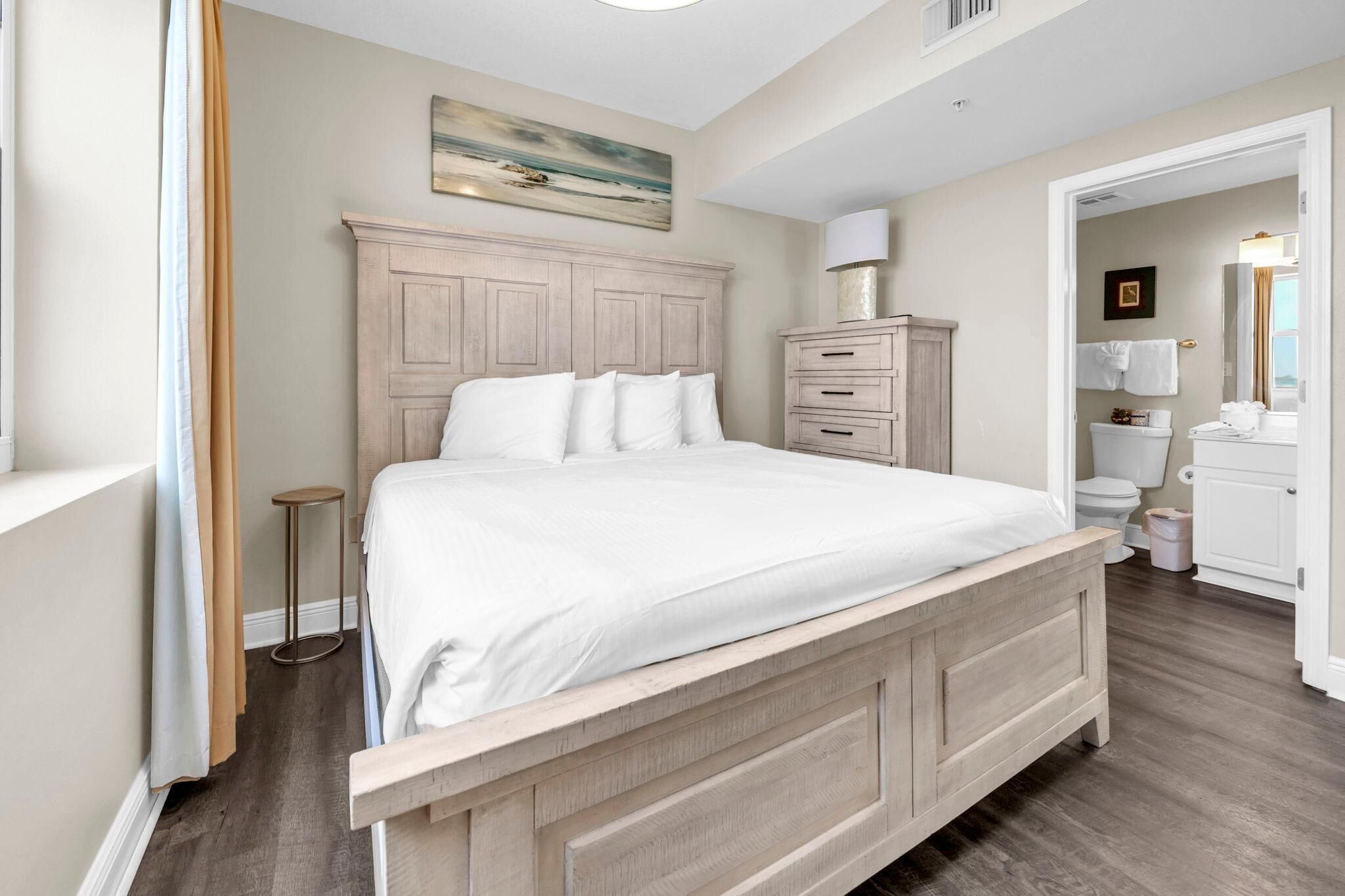 A bedroom with a wooden bed, a nightstand, a dresser, a painting on the wall, and a bathroom visible through an open door.