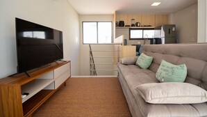 Ein modernes Wohnzimmer mit Flachbildfernseher, einer Couch mit Zierkissen, einer Küchenzeile mit Geräte und einem Fenster, das für natürliches Licht sorgt.