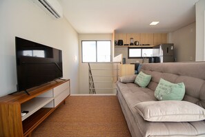 A modern living room with a flat-screen TV, a sofa with decorative pillows, a kitchen area with appliances, and a window providing natural light. - Duplex Vista Mar com Piscina Privativa  (Governador Celso Ramos)