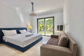 Een slaapkamer met een bed, een bank, een lamp, een televisie en een groot raam met uitzicht op groen.