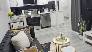 Un salon moderne avec un canapé noir, une petite table ronde et une cuisine en arrière-plan.