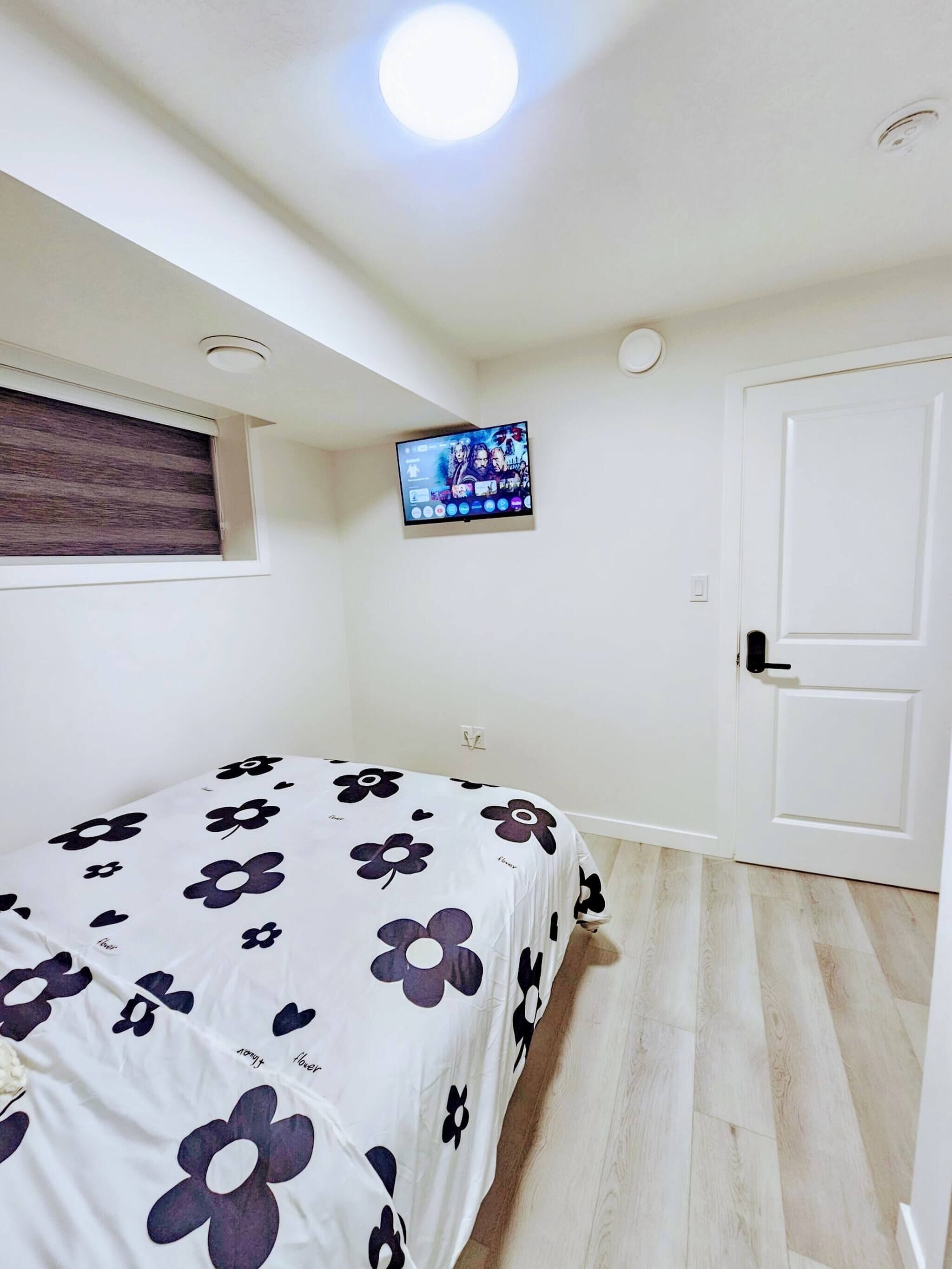 A bedroom with a bed, a television on the wall, a door, and a window with a blind.
