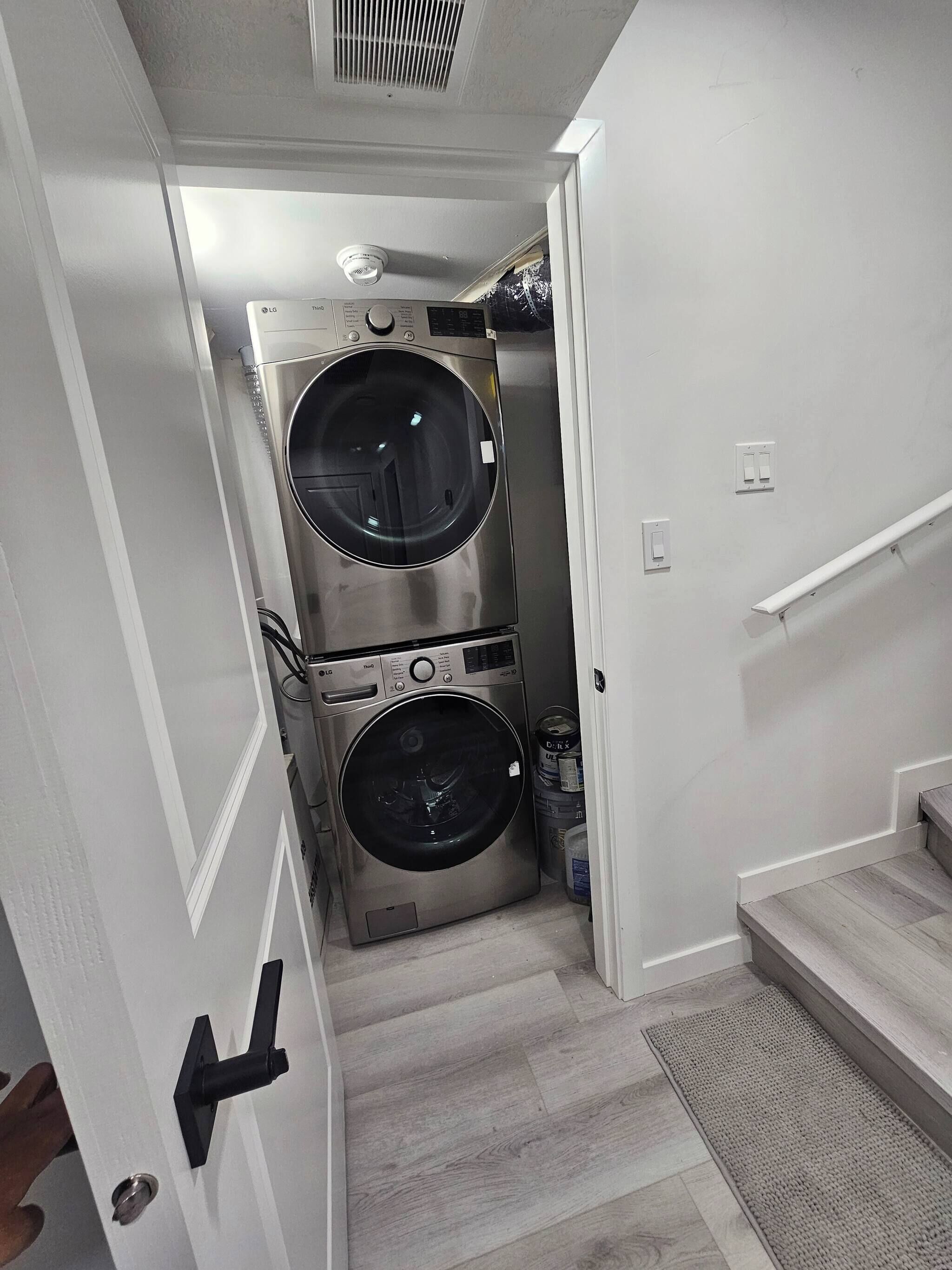 A laundry room with two stacked washing machines, a vent on the ceiling, and a door leading to a staircase.