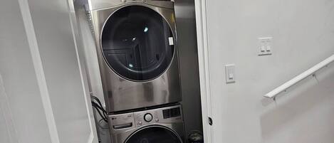A laundry room with two stacked washing machines, a vent on the ceiling, and a door leading to a staircase.