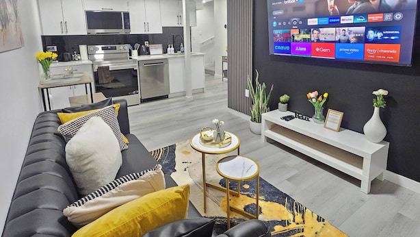 Un salon moderne avec un canapé en cuir noir, un téléviseur à écran plat et une cuisine en arrière-plan.