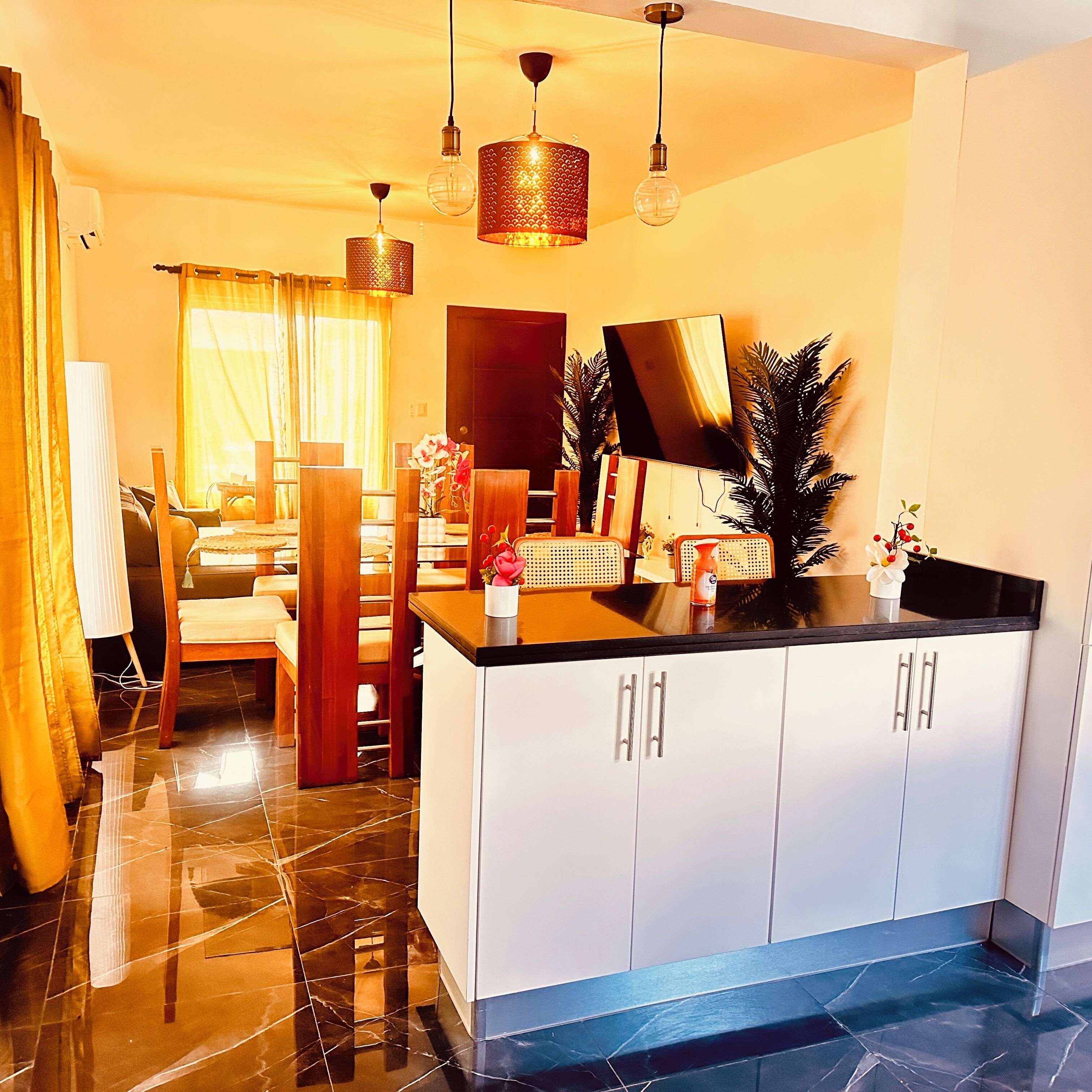 A modern kitchen with white cabinets, a black countertop, and a large island. There are pendant lights above the island and a large mirror above the cabinets.