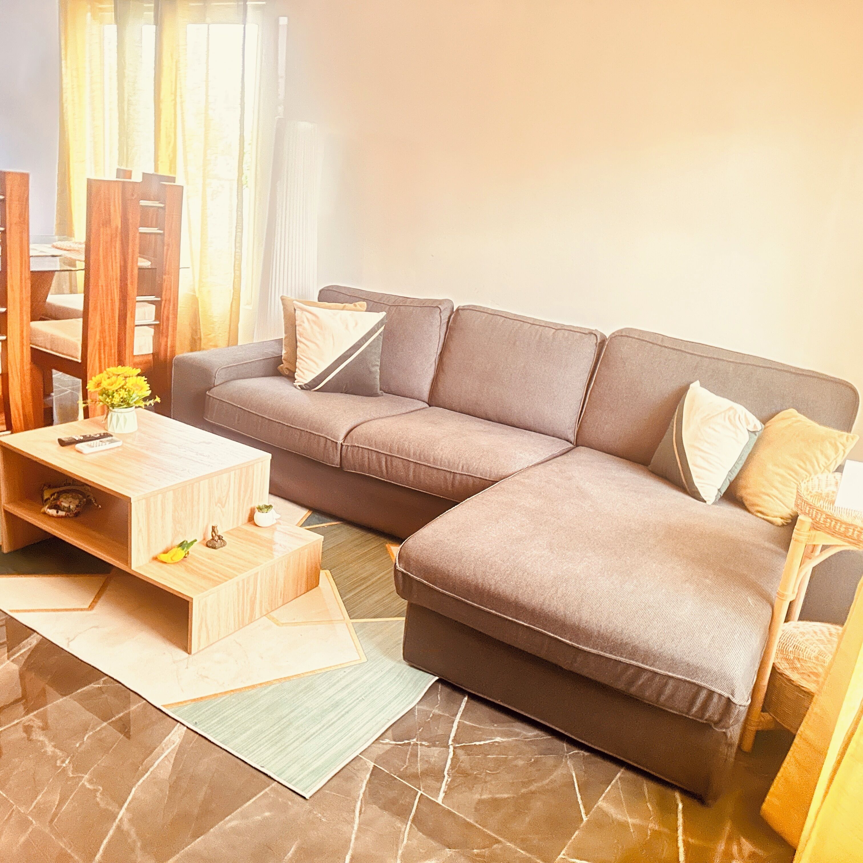 Un salon moderne avec un canapé d’angle, une table basse en bois, une petite table d’appoint et un tapis à motifs.