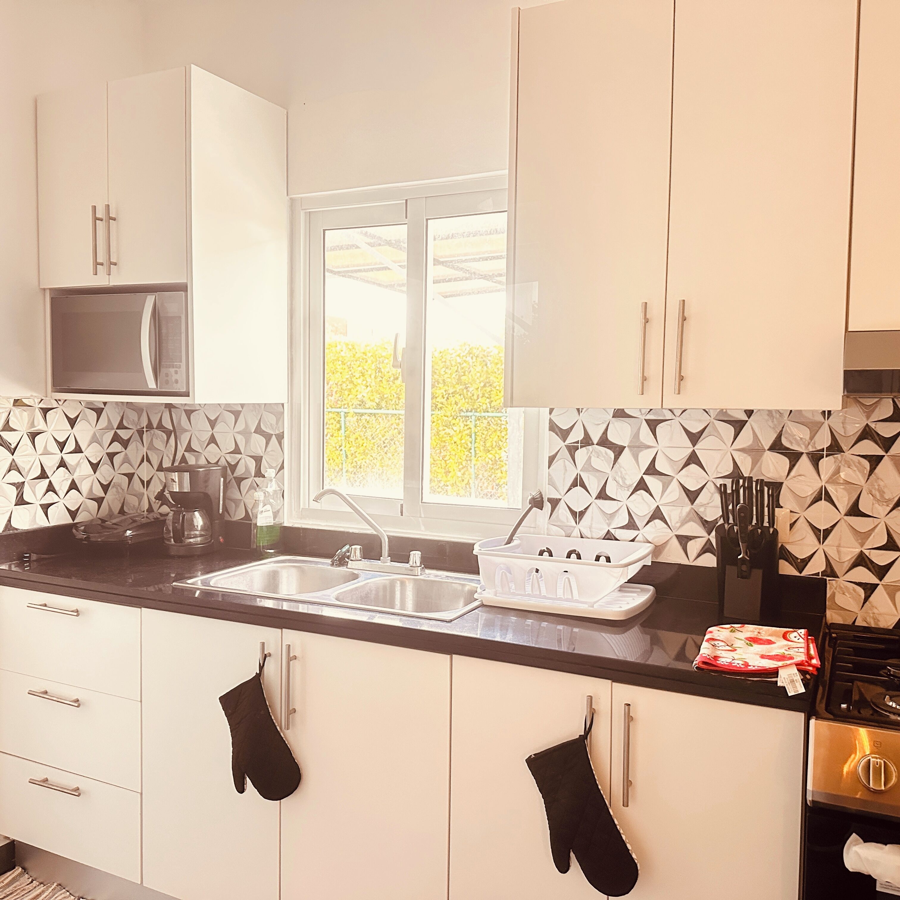 A kitchen with white cabinets, a double sink, a microwave, and a window with a view of greenery.
