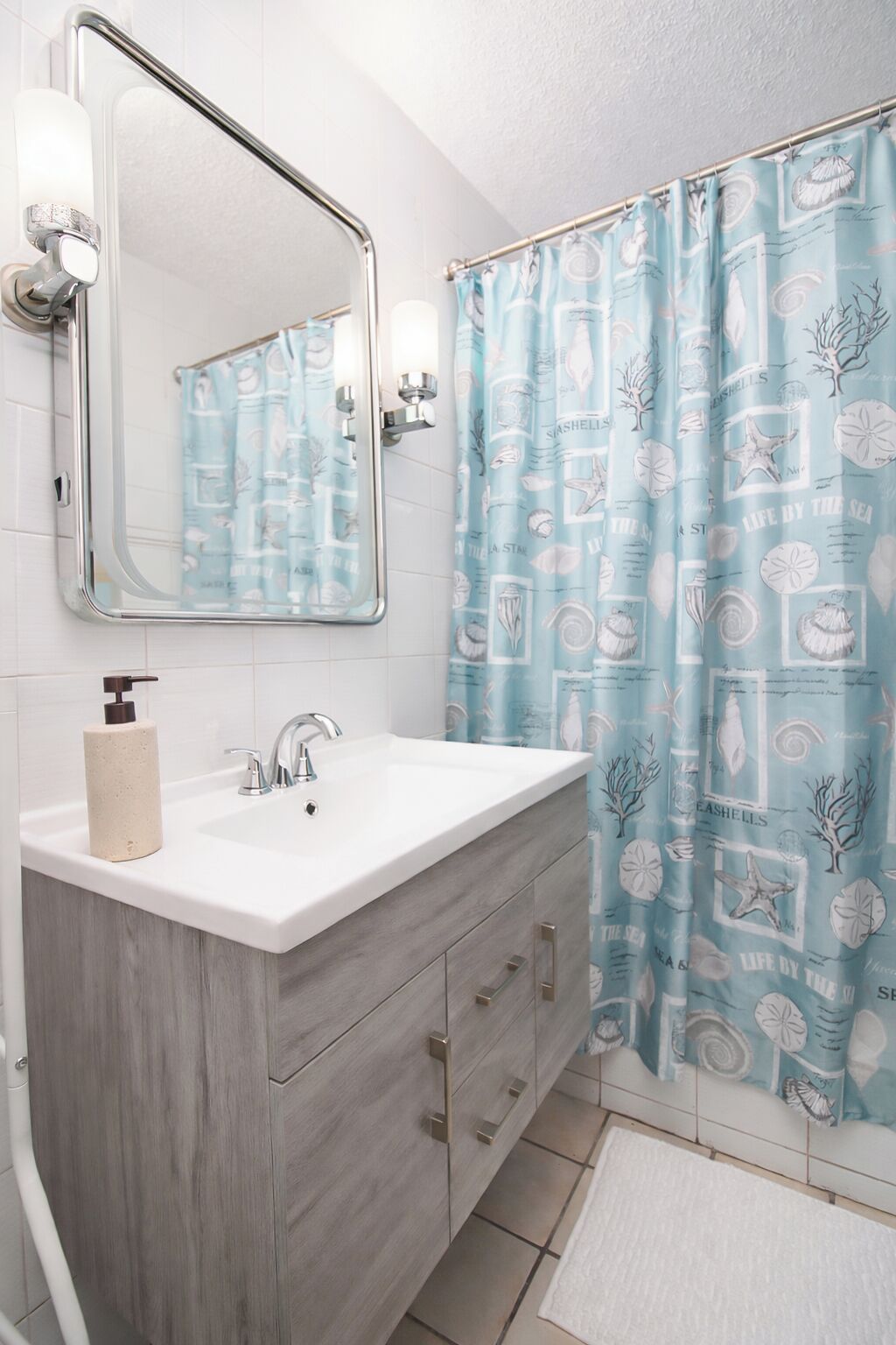 Un baño con lavamanos blanco, un espejo, dispensador de toallas y cortina de ducha con motivos marinos.