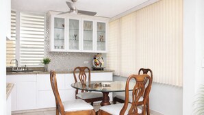 A dining area with a round glass table, four wooden chairs with cushioned seats, a ceiling fan, and a kitchen with white cabinets and a tiled backsplash. - Modern Villa on Puerto Rico's West Coast (Mayagüez)