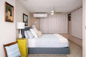 Une chambre à coucher comprenant un lit, des tables de chevet, un ventilateur de plafond, un appareil de climatisation et une fenêtre avec des rideaux.