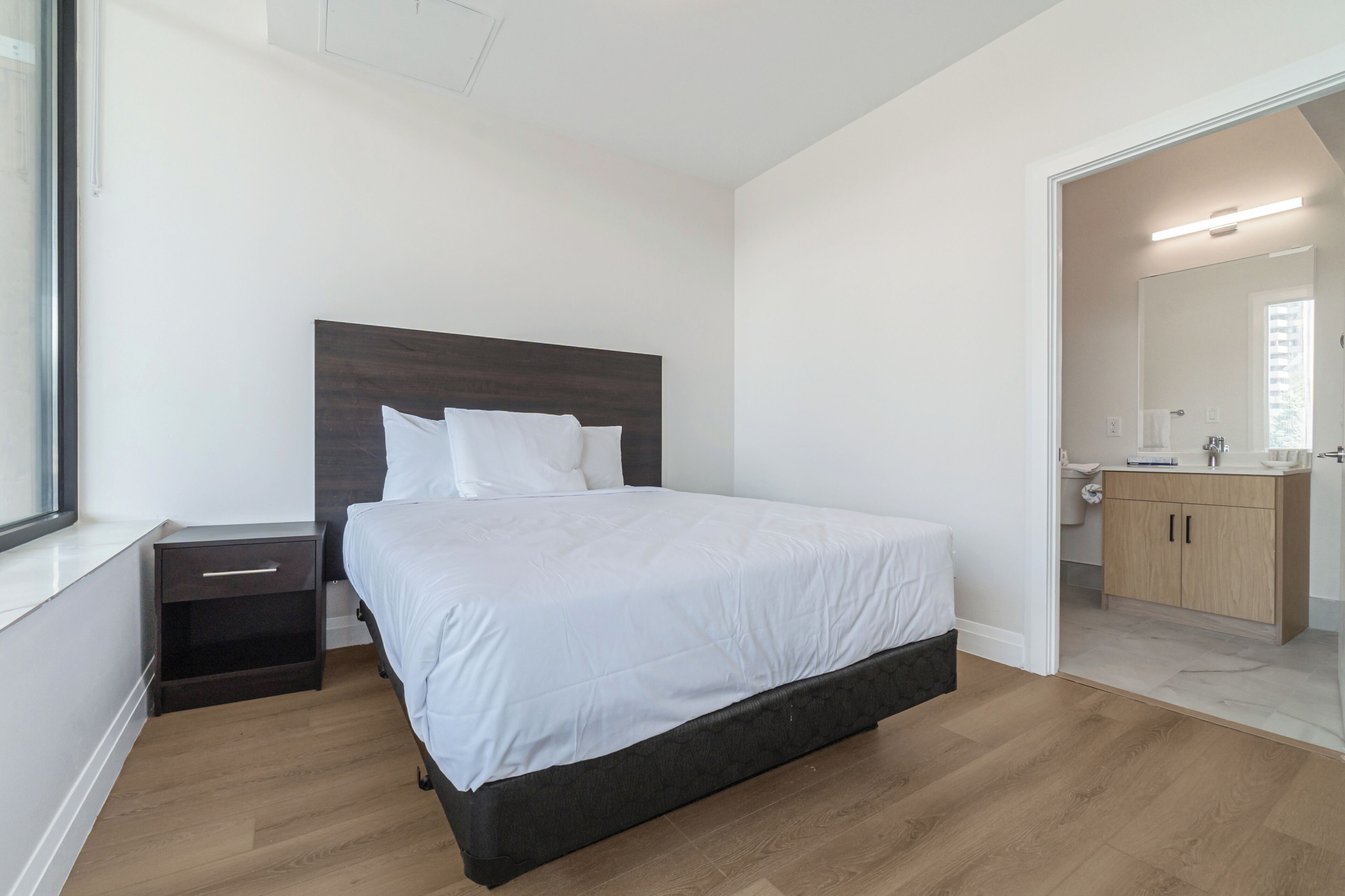 Een slaapkamer met een bed, nachtkastje, houten vloer, een badkamer met spiegel en een kast.